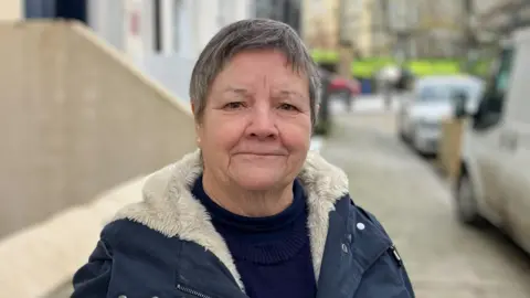 A woman with short cut hair, which is turning grey, is looking at the camera. She has dark eyes and a slight smile on her face. The woman is standing on a street which is blurred in the background. She is wearing a navy, round neck jumper and a navy coat with fur lining. 