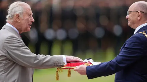 Reuters King Charles III wearing a grey suit and white shirt places his hand on a red cushion held by the Lord Provost of Edinburgh Robert Aldridge, wo wears glasses and a navy blue suit. In the background, lines of soldiers in military uniforms are blurred.