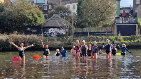 A group of about 13 swimmers, mostly women, standing and waving in the River Wensum. They are in shallow water and some have tow floats. There is some housing in the background.
