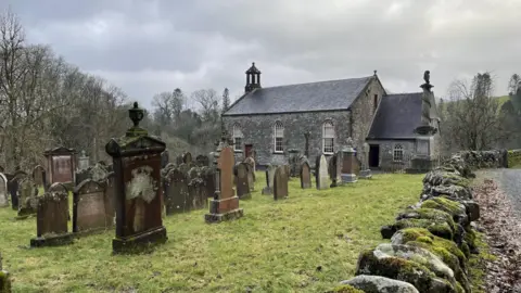 A 19th Century church and cemetery in a countryside setting. There are headstones in the foreground and a dry stone wall at the side.