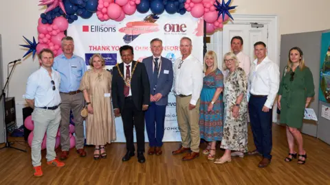 Ellisons Solicitors Ipswich mayor Elango Elavalakan wearing a suit and mayoral chain standing in a line with workers from Ellisons Solicitors at Ipswich Town Hall. Behind them are pink balloons and a screen that reads "Thank you to our sponsors".