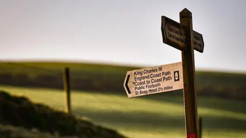 Environment Agency A wooden signpost pointing to King Charles III and Coast to Coast path to St Bees. The signpost is in the foreground with fields in the background, out of focus.
