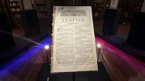 BBC The First Folio on display in a large glass cabinet. It has no cover and the front is a page of The Tempest, slightly yellowed with small black writing on it.