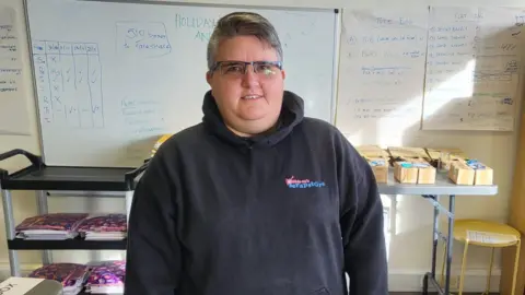 Kelly Wyatt is standing in a room with a whiteboard behind her and a few boxes on a table. She is wearing a black hoodie and glasses. She is smiling.