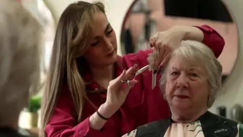 A mature lady is in a hair salon, she is sitting in a chair with a cover over her clothes. A younger woman in red shirt is using scissors and a comb to style to older lady's hair. 