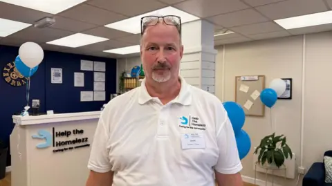 Mark Penn wears a white polo shirt with the Help the Homelezz motif on. He wears his glasses on his forehead. Blue and white baloons have been attached to a desk within the room.
