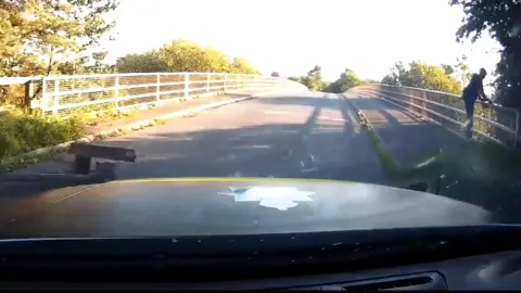 Cumbria Police A silhouette of Daniel Lucas who is running away from a police car, which is shown in the forefront. It is a summer's day and the car is driving on a side road which is crossing the M6 on a bridge. 