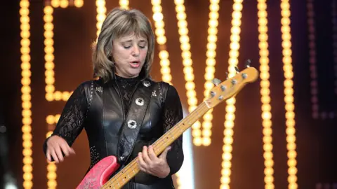 Suzi Quatro wearing a black leather outfit while playing a pink guitar on stage.