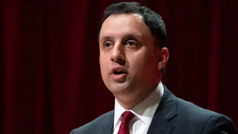 PA Media Anas Sarwar, who has short black hair, wearing a business suit and a red tie. There is a red curtain 