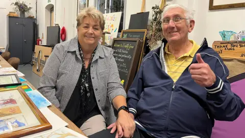 A man and a woman sit in a classroom with chalk boards leaning up against the wall behind them. They are sitting next to a table which is covered in art works, some framed. She has short light brown hair and is wearing a black and white checked shirt over a black top and black and white scarf, she is holding the arm of the man who has short white hair and silver framed glasses. He is wearing a navy jacket over a yellow polo shirt and is doing a thumbs up gesture. Both are smiling.