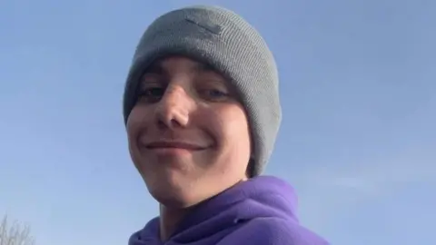 Family photograph Tomasz Oleszak. He is wearing a grey beanie and a purple hoodie. He is smiling and has freckles across his cheeks.