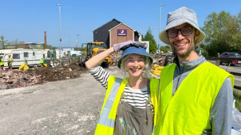 Two people looking at the camera in high-viz jackets. They have bucket hats on and standing on a disused car park. There is rubbish in the background and a big digger. Caravans are on the road and there's a Premier Inn hotel behind.