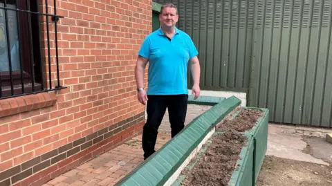 BBC Rick is standing by the empty flower beds, next to a brick wall. He is wearing a light blue polo shirt and has short hair. 