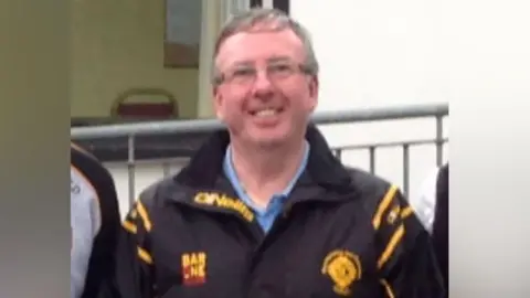 Thomas McKenna smiling. He is wearing glasses and a blue shirt with a black jacket. 