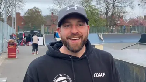 Power Through Sport Johnny Tulip stood in a skate park, wearing a grey cap and a black branded hoodie with the word "Coach" across the chest.