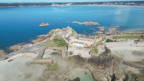 BBC Elizabeth Castle viewed from a drone. There is a large main house at the top of the mound and a castle wall runs around the site. There are larger buildings in a courtyard on the next tier down. There is a small harbour wall to the left of the castle and a beach to the right. There are a few small rocky islets and across the calm water beaches, trees and buildings can be seen.