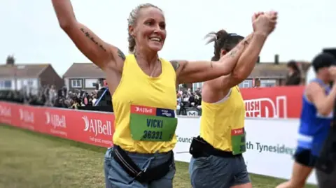 Vikki Hartfree Vikki Hartfree runs with a smile on her face and her arms above her, hand in hand with another female runner, as they cross the finish line of a race. She has blonde hair which has been tied in plaits and she wears a yellow vest top, green shorts and there is a green race number pinned to her vest. 