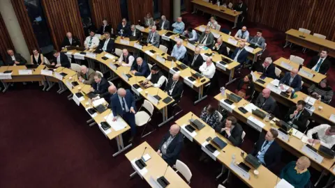 An aerial image of a council chamber, showing councillors sitting at various rows of tables with laptops. 