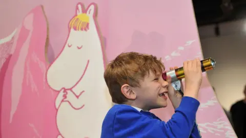 A boy with light brown hair, wearing a blue jumper, looks through a gold and silver telescope. Behind him is a pink board with a white and pink Moomin character on it. 