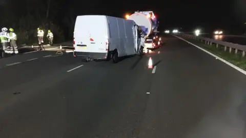 National Highways White van behind a tanker in lane two of a three lane motorway following a crash. It is night. To the left several firefighters are putting up large lights.