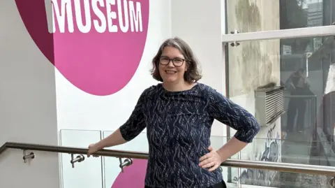 A woman with shoulder length brown and grey hair wearing black-rimmed glasses. She has a navy blue pattered top on and stands in a well-lit corridor. 