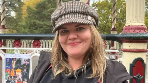 A woman with blonde hair and wearing a cap and a grey top is standing in front of railings. There is a large poster with images on it behind her.