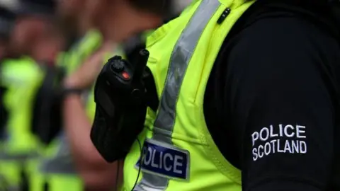 Generic image - Close up of a police officer wearing a black shirt with "Police Scotland" in white stitching on the sleeve and a high visibility vest with "Police" labelling on it and a walky talky strapped to their chest. In the background, other officers are visible but out of focus.