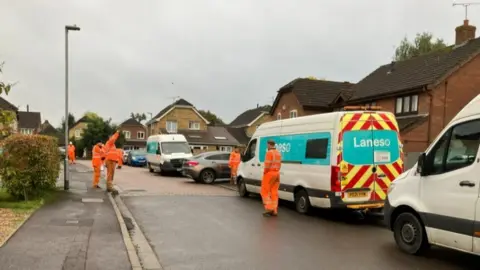 Vans line right hand side of residential street with workers in orange on pavement and in road with homes seen in background