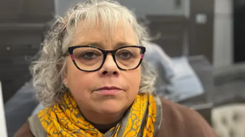 Andy Watson/BBC Maxine Thompson-Curl has short grey hair and is wearing a serious expression. She is dressed in a brown jumper with a gold and silver scarf.  She is wearing black glasses with rainbow-tinted frames.