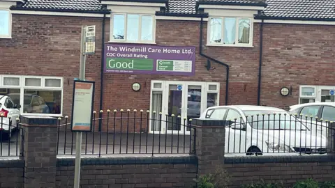 Qays Najm/BBC The outside of a bricked building, with cars parked outside, and a fence and brick wall in the foreground. There is a purple and green-coloured banner pinned to the building reading THE WINDMILL CARE HOME LTD.