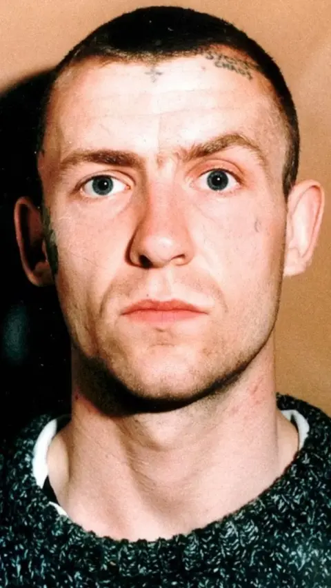 A grainy mugshot of Reginald Wilson at the time of the crime. He has short shaven brown hair and some small facial tattoos on the forehead. He is wearing a grey jumper and looking serious down the camera.