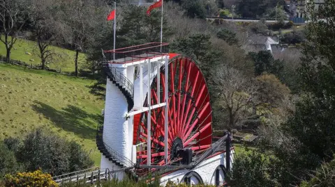 An arial view of the Laxey wheel, a large white structure with a red wheel, and a Manx flag, there are trees in the background.