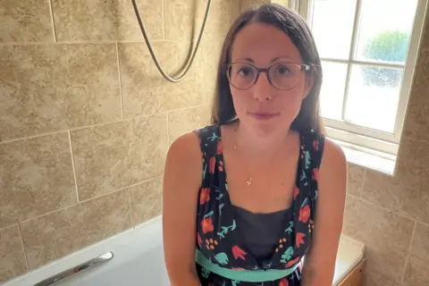 Mousumi Bakshi/BBC A woman with glasses and brown hair, and a floral dress, sitting on the edge of her white bath.