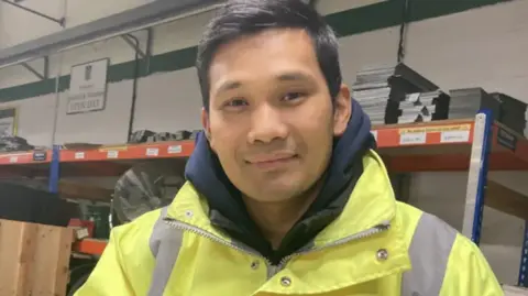 Jo Burn/BBC An image of Tirtha Thapa at the current RBVE factory in Aylesford, Kent. Tirthraj is wearing a high-vis coat and navy hoodie. 