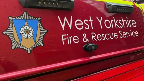 A close up of the side of a red fire service vehicle. Prominently displayed on the vehicle is white text reading “West Yorkshire Fire & Rescue Service”, which stands out clearly against the glossy red paintwork. To the left of the text, an official service crest is visible, featuring a white rose emblem set within a shield and surrounded by a starburst-style badge.