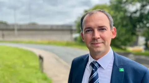 Chris Wood, Plymouth City Council member, in a blue shirt, blue striped tie and blue suit jacket with a green Plymouth City Council badge.