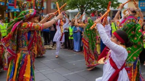 A previous Festival of Chariots event in Leicester with a group of people dancing with brightly-coloured clothes and making music with yellow and red-striped sticks
