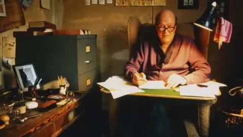 Jan Baldwin The author Roald Dahl wearing a cardigan and shirt in his writing hut. There is a lamp over his left shoulder. He is surrounded by a filing cabinet and another desk which has various items on it such as a brush and a pipe.