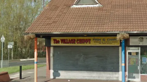 General view of the Village Chippy. It is a unit on a small parade of shops. Its shutters are down in the image.