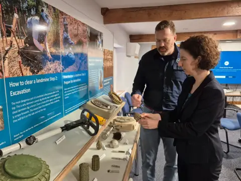 UK Government International Development Minister Annaliese Dodds is shown around the Halo Trust landmine clearance charity headquarters by a man with a beard in a blue shirt as they look at metal detectors and other demining equipment