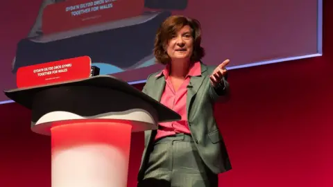 Matthew Horwood Eluned Morgan stood at a lecturn at party conference, with a red background behind her, wearing a green jacket and trousers and a red shirt
