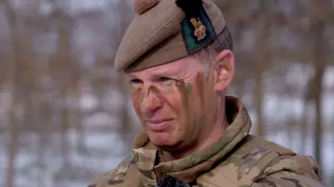 Brigadier Andy Watson, who is commanding the British contribution to the Nato exercise, is shown wearing an army uniform with green and brown camouflage paint on his face. He is also wearing a green and brown military beret hat 