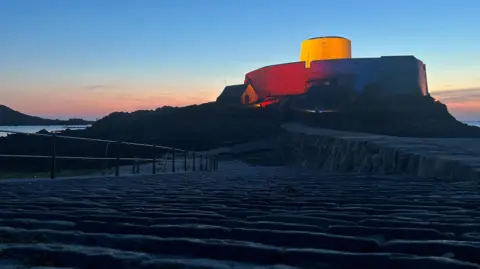 A view of Fort Grey lit up in red, white and blue. The sun is setting in the background.