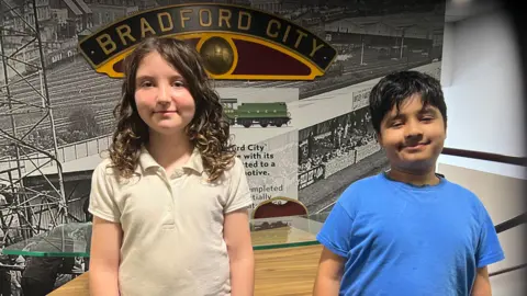 Aisha Iqbal/BBC Two children stand in front of a photo feature wall showcasing the history of Bradford City. The girl has long, mousy brown and wavy hair and wears a white polo shirt. The boy is shorter, has short black hair and wears a blue T-shirt.