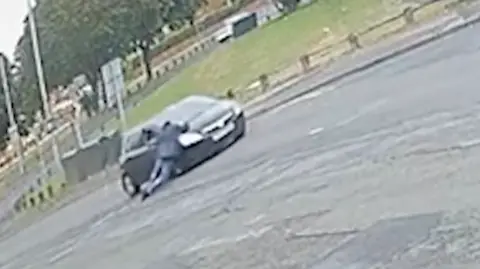 A man is being dragged along the road while he holds on to the bonnet and driver-side door of a black hatchback car.