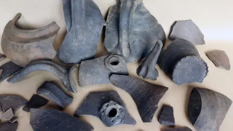 Albion Archaeology/Central Bedfordshire Council A selection of dark broken pottery, some with handles and basic decoration, are laid out on a light-coloured surface .