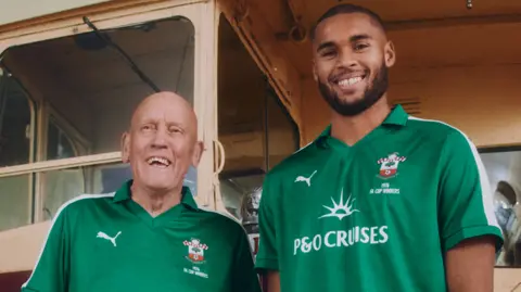 Southampton Football Club Ian Turner and Gavin Bazunu where the club's new green commemorative goalkeeper shirts while standing in front of the original celebration bus. The shirts feature a P&O Cruises logo and the words '1976 FA Cup winners'. Both men are grinning widely. 
