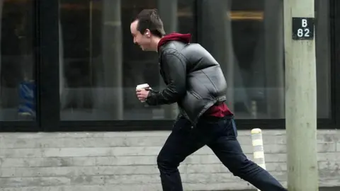 Getty Images A man wearing a black coat and jeans, carrying a coffee cup, struggling to cross a road during a strong wind
