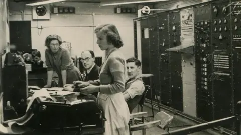 A black and white photo of four people, two men and two women, stood and sat around a desk looking at instruments.
