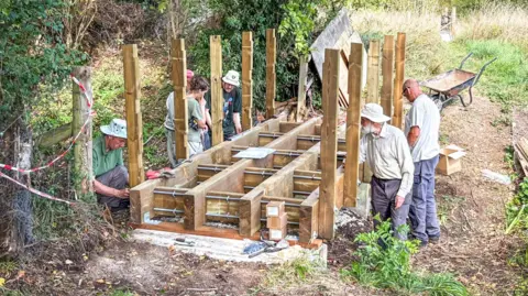 A group of volunteers builds a wooden footbridge structure in an outdoor rural area. Several upright timber posts are fixed into the ground, forming the early framework of the bridge. 